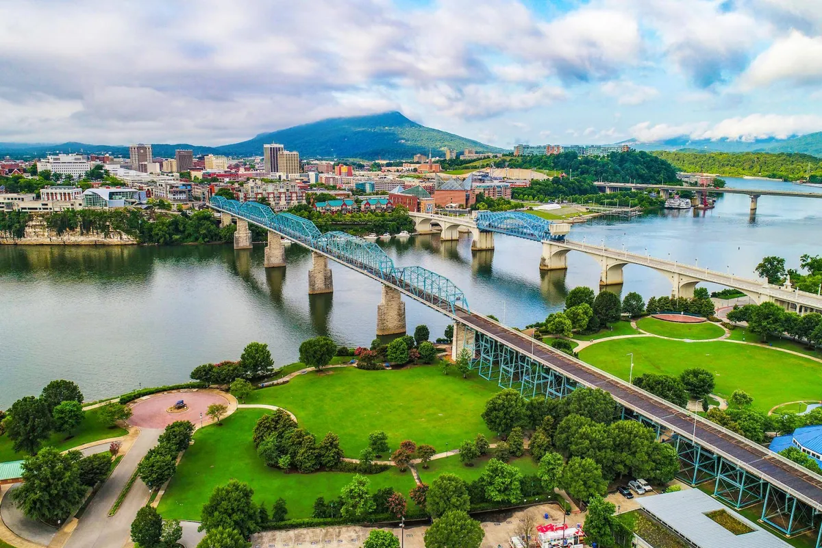 This image presents a sweeping view of Chattanooga's riverfront, featuring bridges, a city skyline, lush green parks, and nearby mountains. The scene captures both natural beauty and urban vibrancy, with visible walking and cycling areas, as well as open spaces ideal for festivals and family outings.