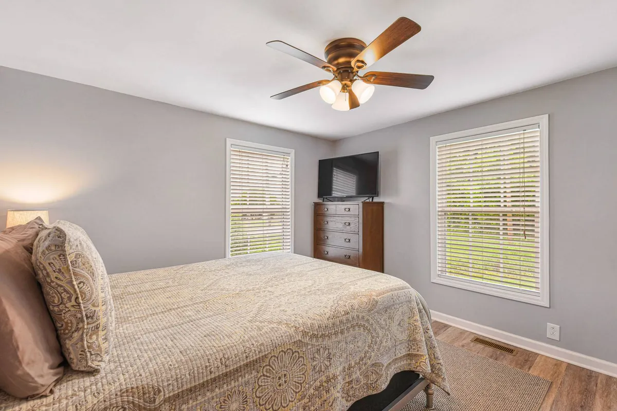 A bright queen bedroom featuring a comfortable bed with plush bedding, a spacious dresser, large windows letting in natural light, and a ceiling fan above. The setup includes a Smart TV positioned for easy viewing from the bed, with a tranquil, uncluttered décor that invites relaxation.