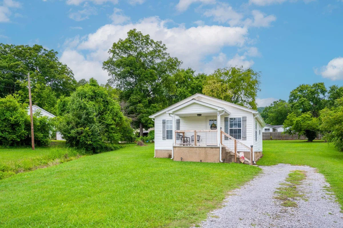 This image showcases the front exterior of Tennessee Delight 2, featuring a welcoming porch, lush green lawn, and a gravel driveway. The spacious and well-maintained yard is visible, perfect for outdoor relaxation or playtime with pets.