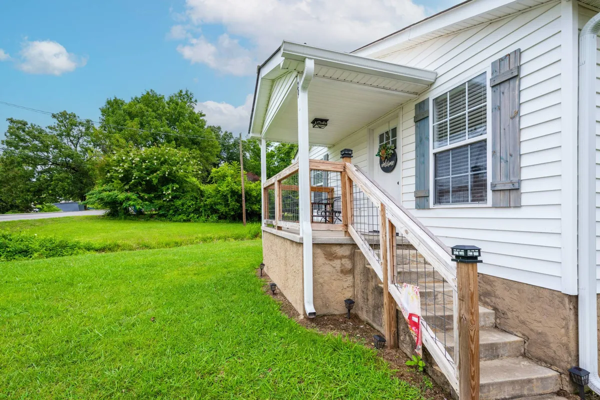 This image features the home's inviting wooden steps and front porch surrounded by a green lawn and mature trees. The scene naturally draws the eye toward the tranquil porch space, perfect for relaxing with a cup of coffee or unwinding after a day of adventure. The soft lighting hints at a peaceful sunset, and the overall mood is one of quiet comfort and welcoming atmosphere.