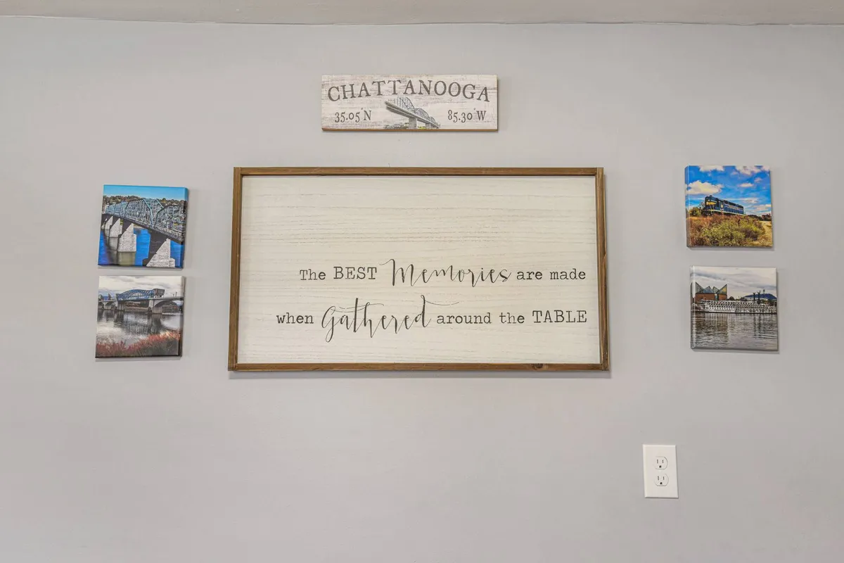 A thoughtfully styled interior scene with a framed sign, photographs of Chattanooga, and a wooden sign with coordinates. The dining area is visible, suggesting a space meant for shared meals and meaningful conversation.