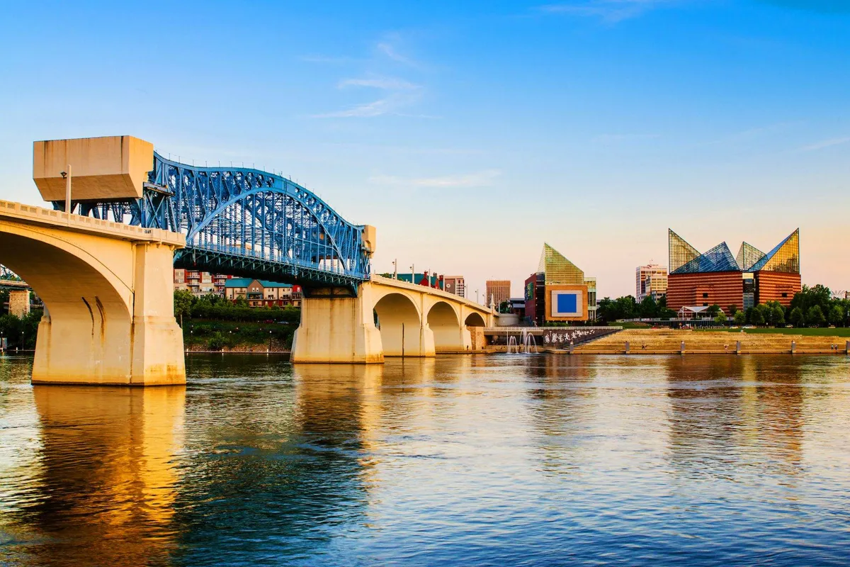 A captivating view of the blue bridge stretching over the river, flanked by modern buildings and embraced by serene water and sky. The scene encapsulates the essence of Chattanooga’s Riverwalk area, inviting viewers to imagine walking or biking along the river and soaking up local art, culture, and natural beauty.