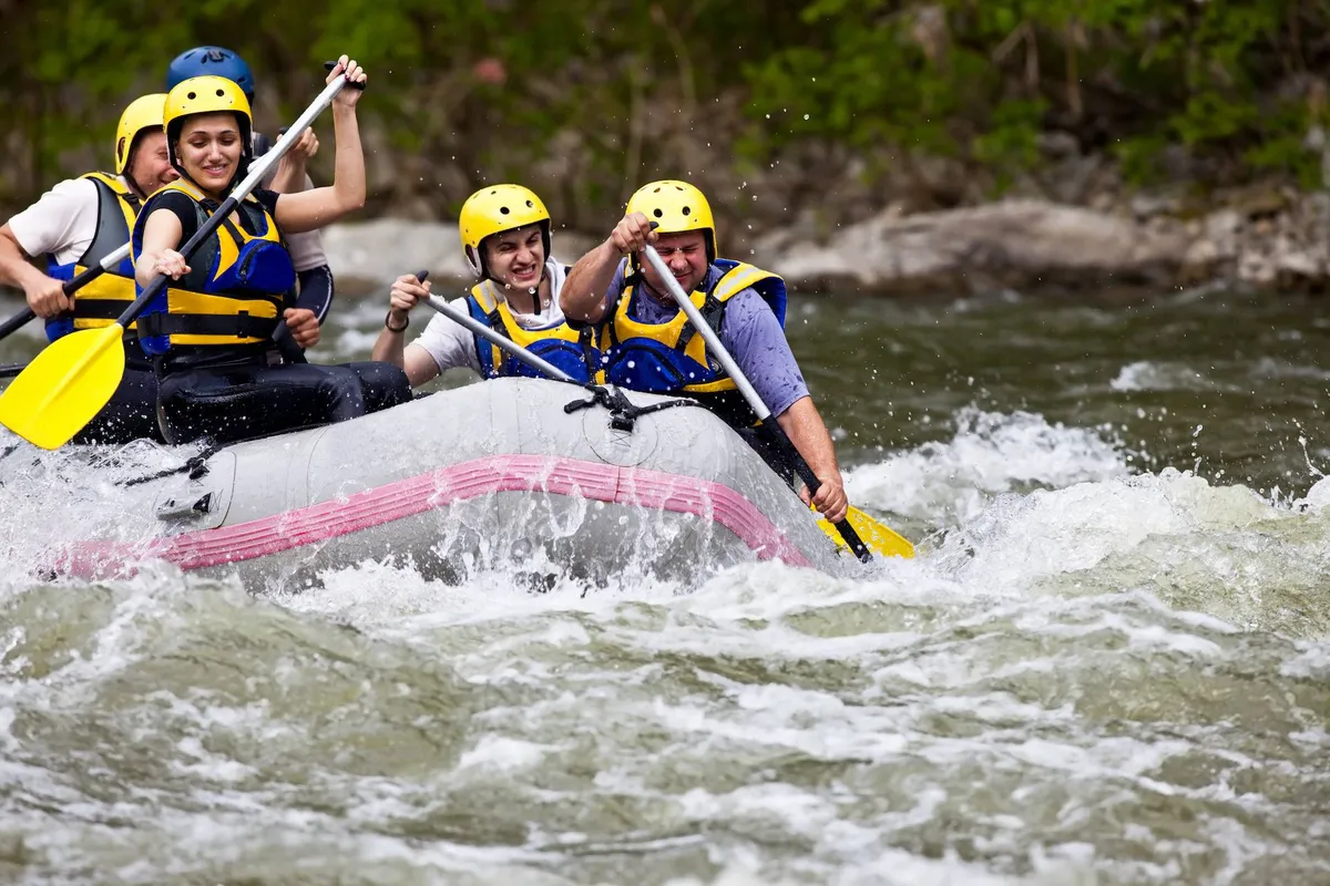 A group of people navigate a raft through rushing river rapids, paddles raised in unison as they tackle the waves together. The backdrop features lush greenery and the powerful flow of the river, highlighting the excitement and energy of the outdoor adventure.