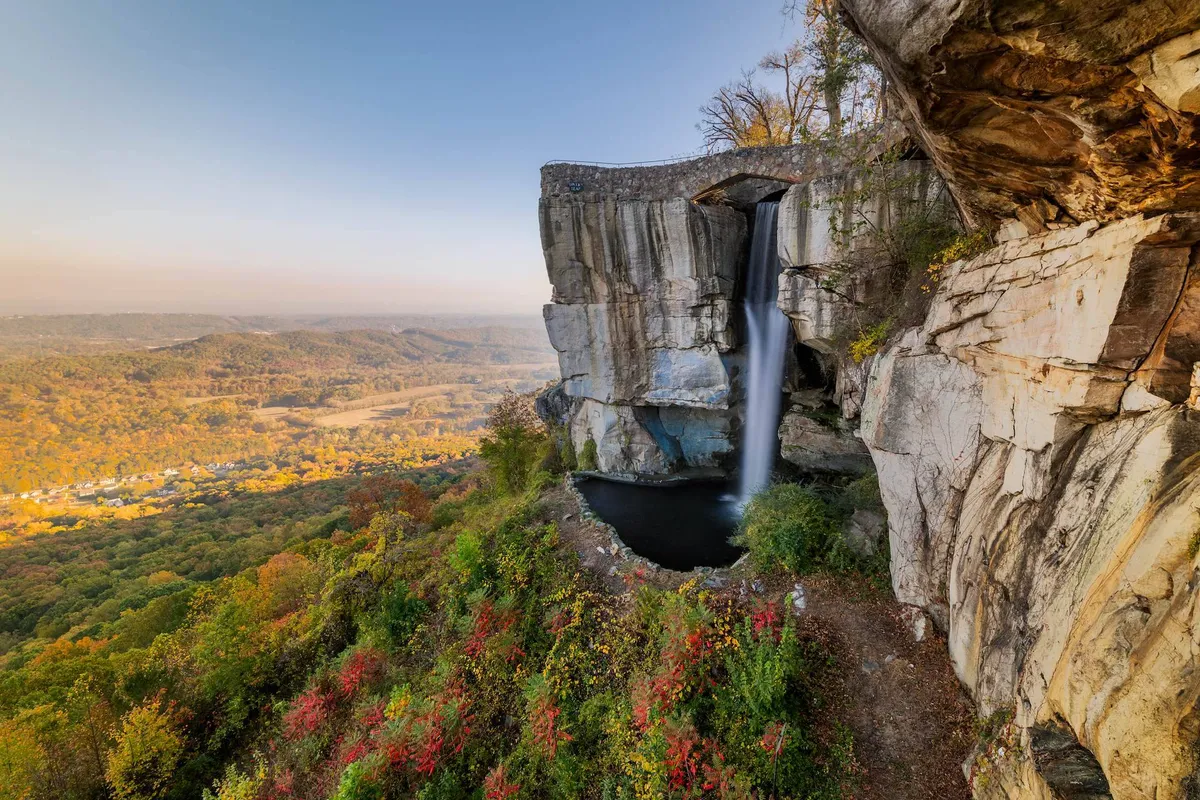 A picturesque waterfall cascades over rock formations into a lush valley surrounded by vibrant autumn foliage. The image hints at hiking trails and picnic spots, evoking a sense of discovery and natural beauty that mirrors the nearby attractions like Rock City and Ruby Falls.