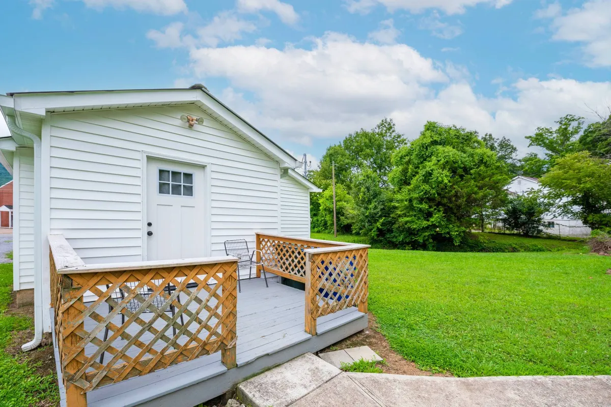 A peaceful exterior shot of a white wooden house with a spacious porch, deck railing, and a green lawn surrounded by trees. The scene suggests opportunities for morning strolls, sunset views, and stargazing with pets.