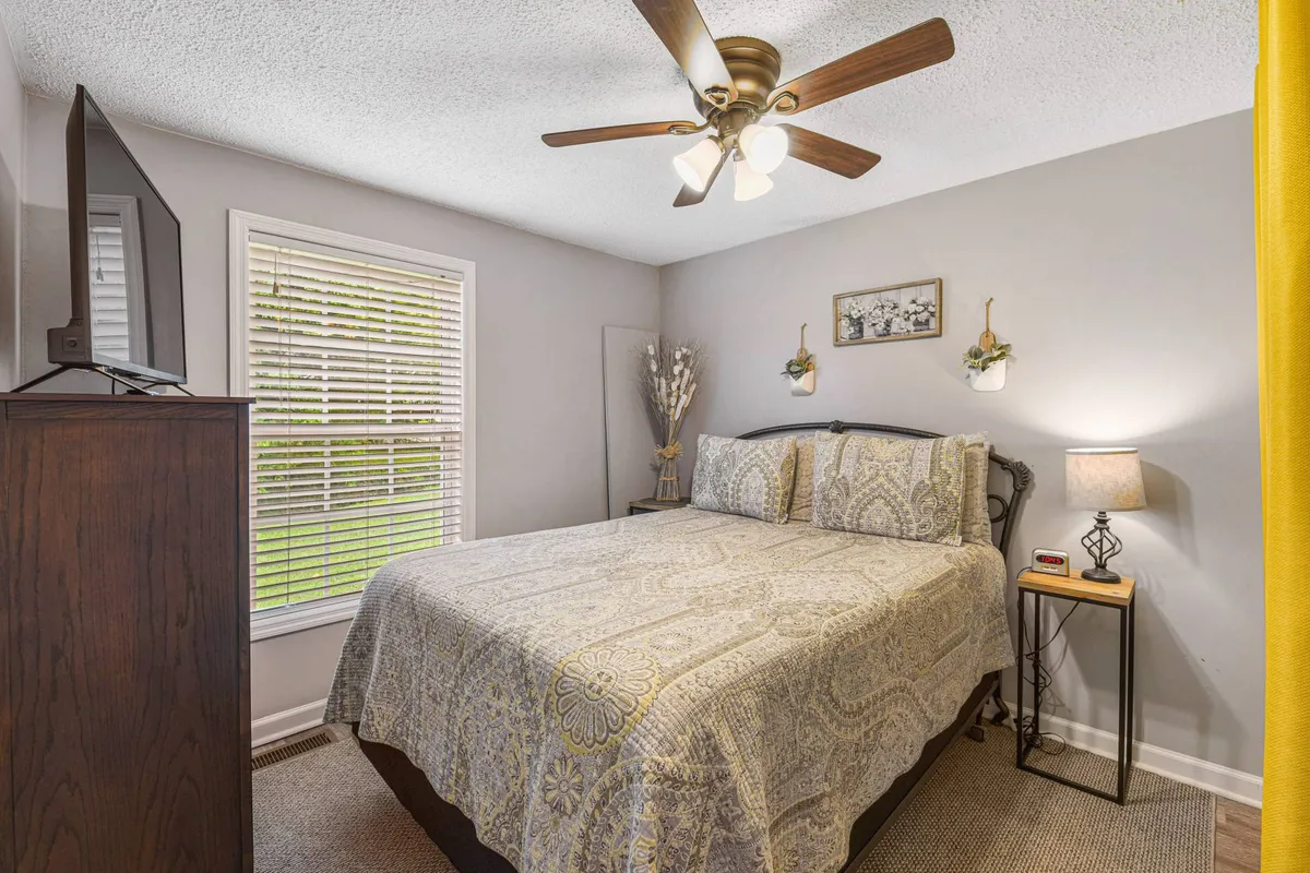 A cozy queen bedroom decorated with patterned bedding and soft lighting from a bedside lamp. The room features a Smart TV, a window with blinds, and a nightstand, creating a welcoming environment ideal for unwinding at the end of the day.
